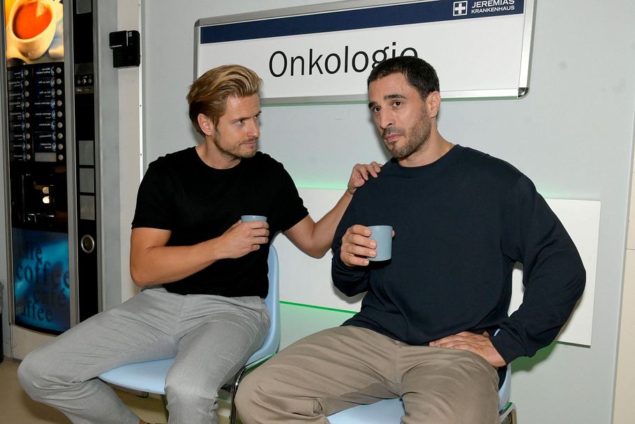 GZSZ-Szene: Philip sitzt neben Tobias in der Klinik vor dem Schild mit der Aufschrift Onkologie