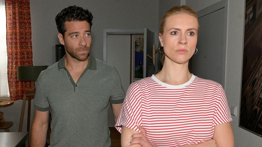 GZSZ-Szene Zoe steht mit verschränkten Armen in ihrer Wohnung, Carlos steht hinter ihr und schaut sie nervös an