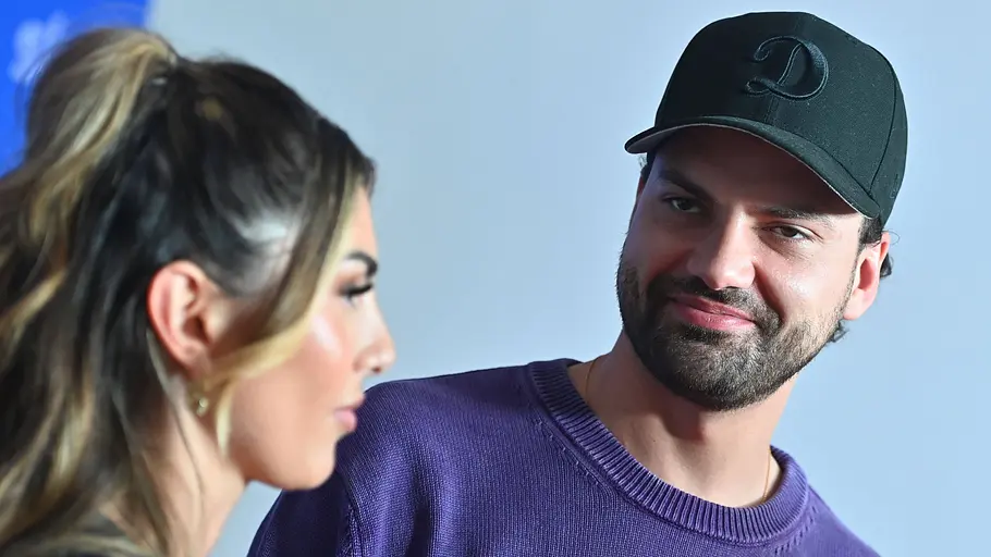 „Yeliz & Jimi – We Are Family?!“-Ärger Jimi Blue Ochsenknecht trägt eine schwarze Kappe und einen violetten Pullover und blickt zu Yeliz Koc, die im Profil zu sehen ist.