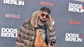 Haftbefehl trägt Sonnenbrille, Pelzjacke und eine braune Umhängetasche vor einer Wand mit Netflix- und „Dogs of Berlin“-Logos. - Foto: IMAGO / Photopress Müller Haftbefehl trägt Sonnenbrille, Pelzjacke und eine braune Umhängetasche vor einer Wand mit Netflix- und „Dogs of Berlin“-Logos. - Foto: IMAGO / Photopress Müller