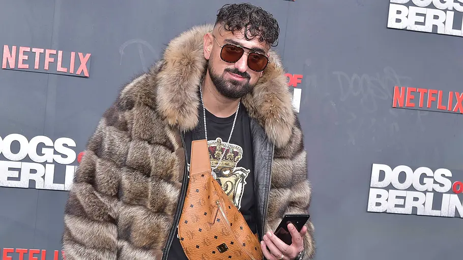 haftbefehl rückfall drogen Haftbefehl trägt Sonnenbrille, Pelzjacke und eine braune Umhängetasche vor einer Wand mit Netflix- und „Dogs of Berlin“-Logos.