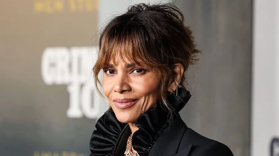 Halle Berry trägt einen schwarzen Blazer mit hohem Rüschenkragen und goldenen Schmuck. Ihr Haar ist hochgesteckt.
