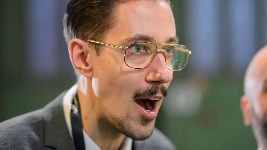 HandOfBlood trägt eine goldfarbene Brille und ein graues Sakko. Er blickt mit offenem Mund nach rechts.