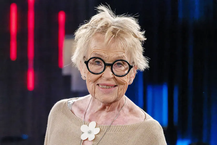 Hannelore Becker Hannelore Becker lächelt mit zerzausten kurzen blonden Haaren und markanter runder schwarzer Brille in die Kamera. Sie trägt einen beigen Pullover mit einer großen weißen Blumenkette, im Hintergrund sind unscharfe Lichtelemente eines TV-Studios zu sehen.