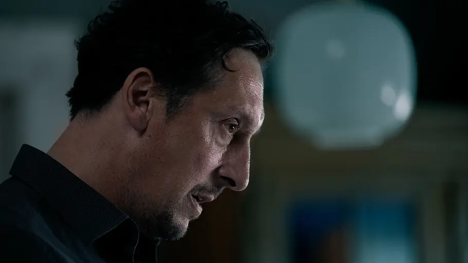 Hans Löw als Robert Lenzen in „Die Falle“. Hans Löw als Robert Lenzen mit dunklen Haaren und Kinnbart blickt im Halbprofil nach rechts. Er trägt ein dunkles Hemd vor unscharfem Hintergrund.