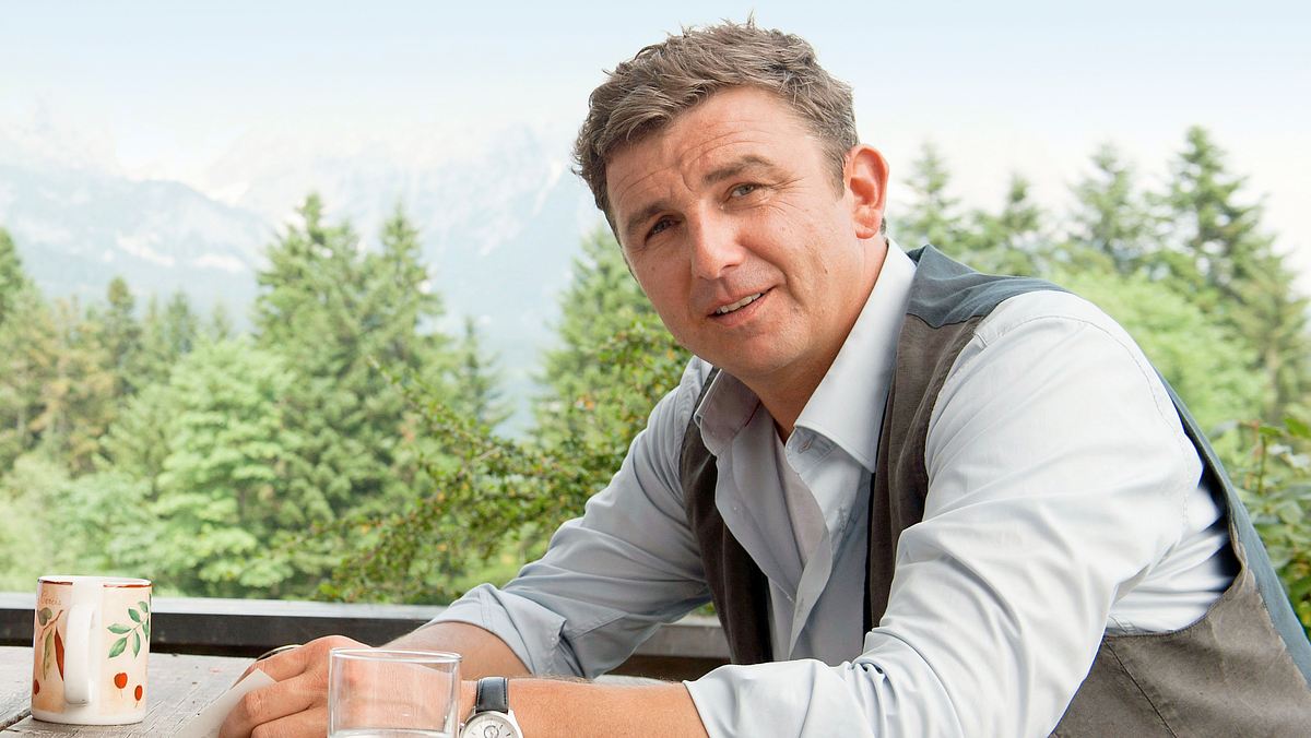 Hans Sigl in „Der Bergdoktor“. Hans Sigl sitzt in Hemd und Weste in „Der Bergdoktor“ draußen an einem Tisch. Hinter ihm sieht man Bäume, weiter in der Ferne sind Berge zu erkennen.