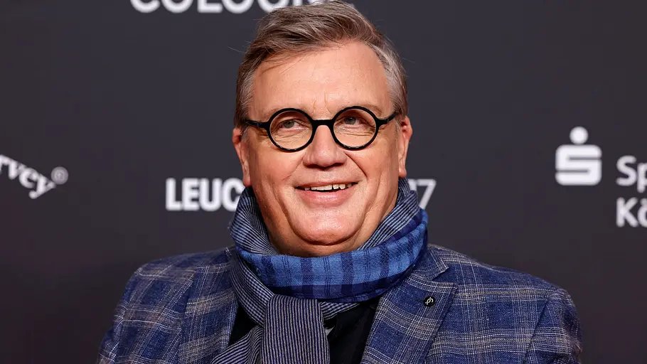Hape Kerkeling lächelt bei einem öffentlichen Auftritt mit Brille, Schal und Sakko vor dunklem Hintergrund.