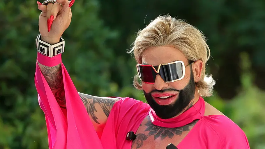 Harald Glööckler soll dabei sein. Harald Glööckler winkt mit pinkem Outfit und Sonnenbrille.