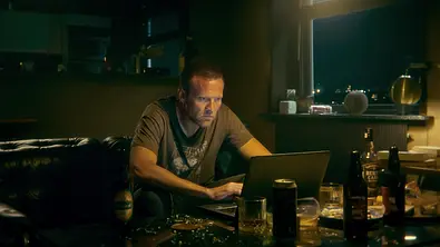 Detektiv Harry Hole sitzt in T-Shirt an seinem Schreibtisch und schaut gebannt auf seinen Laptop - Foto: Netflix