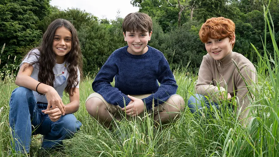Herimine, Harry und Ron-Darsteller Arabella Stanton, Dominic McLaughlin, and Alastair Stout sitzen in der Hocke in einem Feld