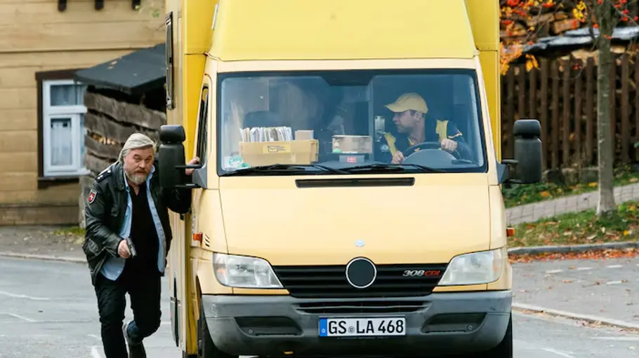 Frank Koops ermittelt in „Harter Brocken: Der Geheimcode“ Frank Koops läuft neben einem gelben Lieferwagen her, während der Fahrer am Steuer sitzt.