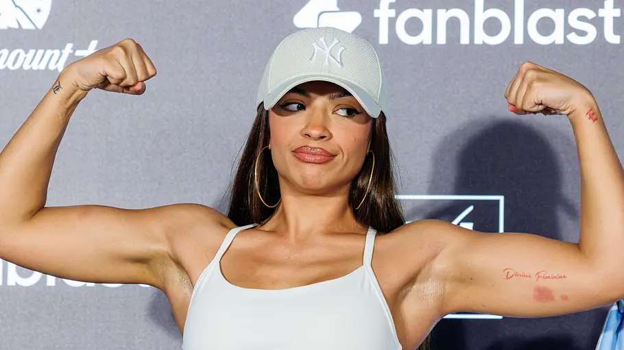 Hati Suarez Fame Fighting Hati Suarez posiert mit erhobenen Fäusten und angespannten Armen vor einer Logowand. Sie trägt eine helle Kappe und ein helles Top.
