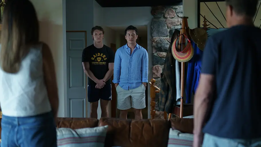 Ilya und Shane stehen nebeneinander in einem Hausflur und blicken ernst nach vorne. Einer trägt ein schwarzes T-Shirt und Shorts, der andere ein hellblaues Hemd und helle Shorts. Im Vordergrund sind unscharf Rückenansichten einer Frau und eines Mannes zu sehen, die vor dem Sofa im Wohnzimmer stehen. Die Szene wirkt angespannt oder ernst.