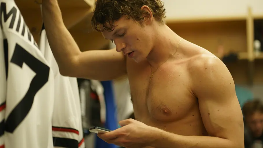 Ilya steht oberkörperfrei in einer Umkleidekabine und stützt sich mit einem Arm an einem Spind ab. In der anderen Hand hält er ein Smartphone und blickt nachdenklich darauf. Neben ihm hängt ein weißes Eishockey-Trikot mit der Nummer 17.