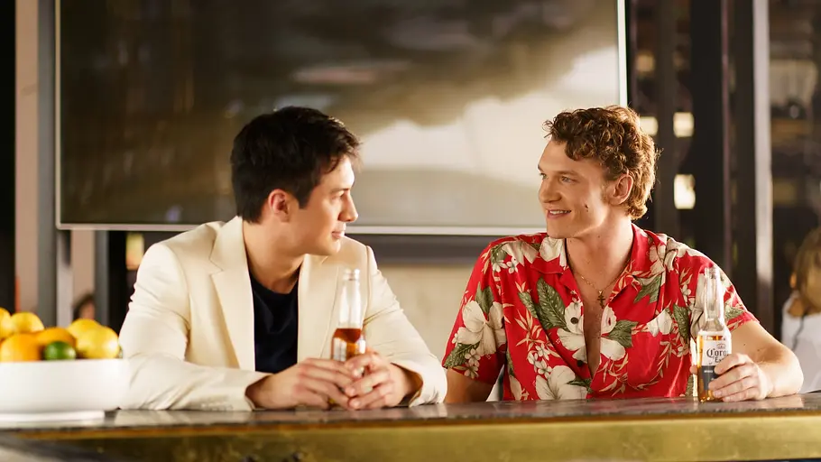 Ilya und Shane sitzen an einer Bar und unterhalten sich lächelnd. Einer trägt ein weißes Sakko, der andere ein rotes Hawaii-Hemd mit Blumenmuster. Beide haben jeweils eine Bierflasche in der Hand. Die Stimmung ist entspannt und freundlich. Im Vordergrund steht eine Schale mit Zitrusfrüchten.
