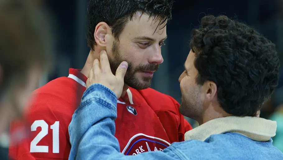 Zwei Männer stehen sich nah gegenüber. Der eine trägt ein rotes Eishockey-Trikot mit der Nummer 21, der andere eine blaue Jeansjacke. Der Mann in der Jeansjacke hält das Gesicht des Mannes im Trikot liebevoll und sie schauen sich intensiv in die Augen. Die Szene wirkt emotional und innig.