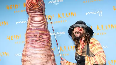 Heidi Klum steht im riesigen Wurmkostüm neben einem Mann mit Angel, schwarzem Hut, Wathose und blutiger Gesichtsschminke. - Foto: IMAGO / Cover-Images