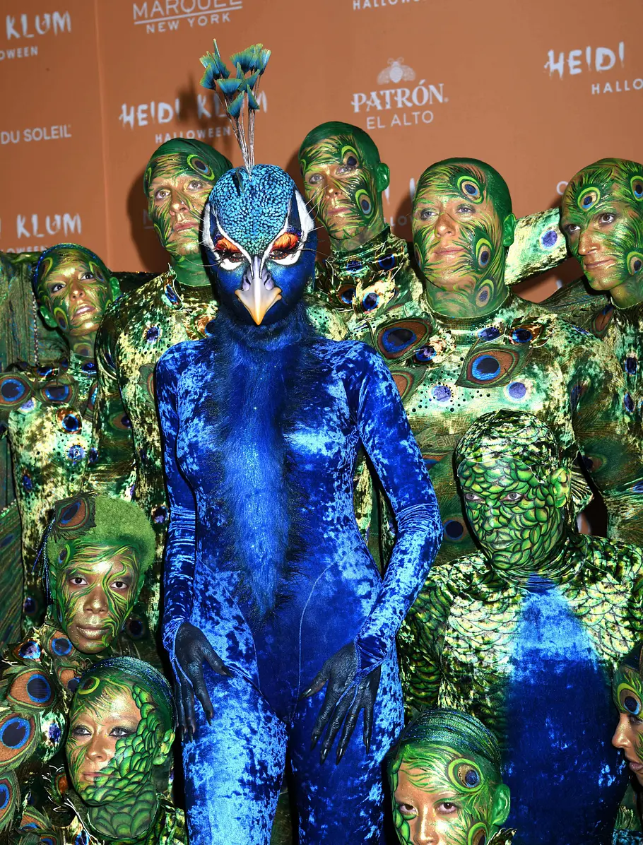 Heidi Klum zu Halloween 2023 als Pfau Heidi Klum posiert an Halloween 2023 im blauen Pfauenkostüm mit Schnabelmaske zwischen grün-golden kostümierten und geschminkten Cirque-du-Soleil-Artisten.