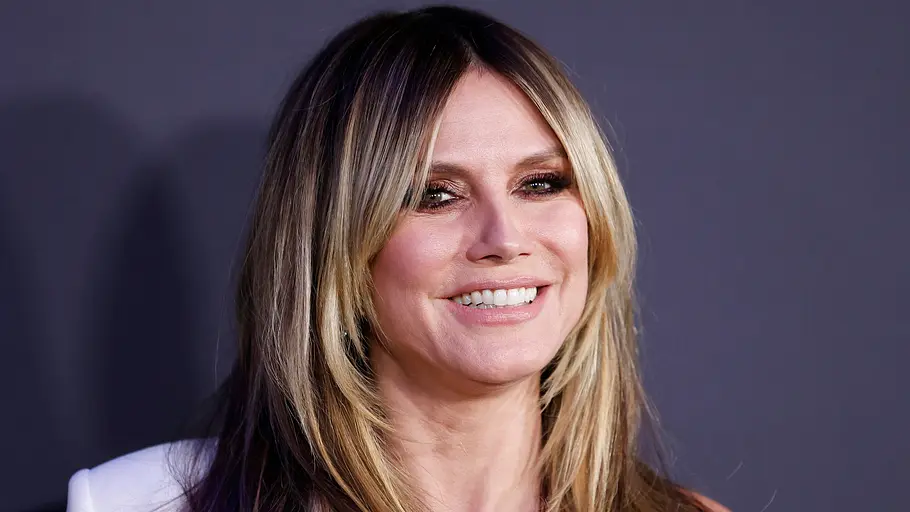 Heidi Klum Heidi Klum steht vor einem dunklen, violetten Hintergrund und grinst in die Kamera.