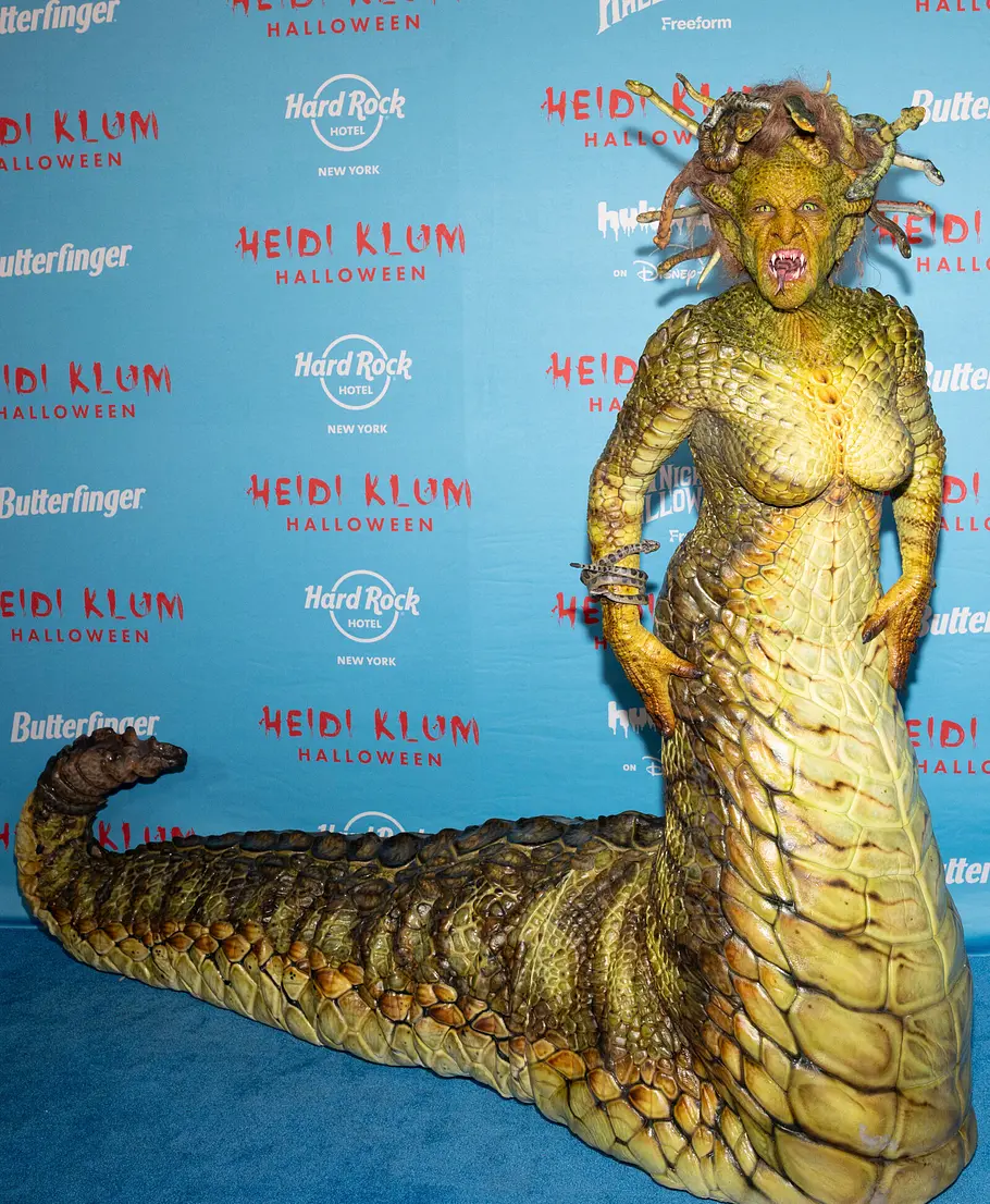 Heidi Klum zu Halloween 2025 als Medusa Heidi Klum posiert an Halloween 2025 in einem goldgrünen Schlangenkostüm mit langem Schwanz und Schlangenkopfputz vor einer blauen Logowand.