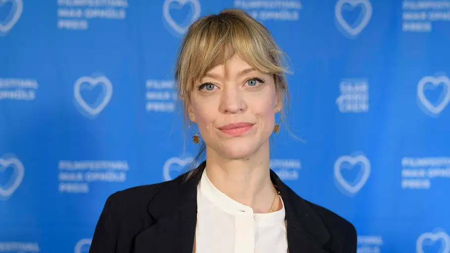 Heike Makatsch trägt einen schwarzen Blazer über einer weißen Bluse vor einer blauen Wand mit Schriftzügen und Herzlogos.