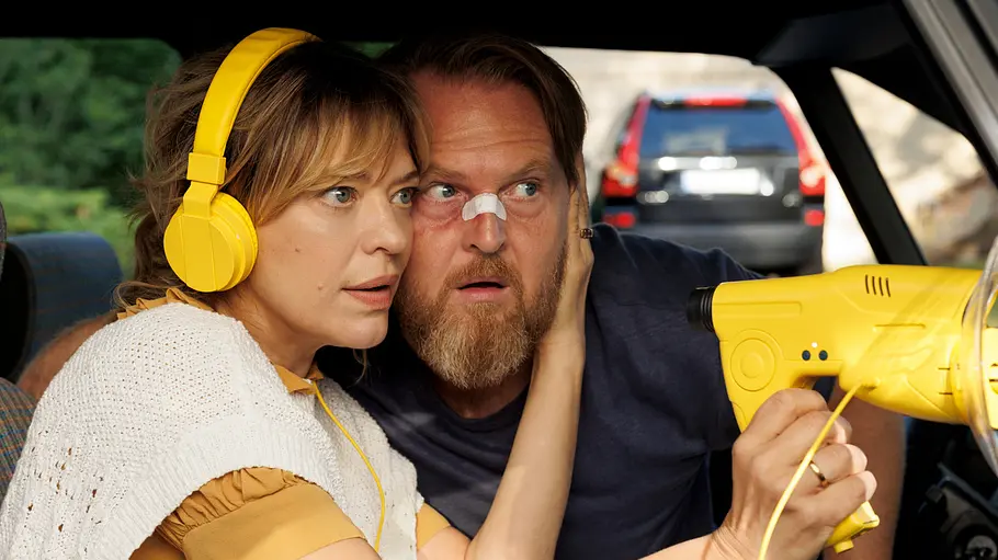 Heike Makatsch und Axel Stein in Where's Wanda? Heike Makatsch trägt gelbe Kopfhörer und hält ein gelbes Richtmikrofon. Axel Stein sitzt mit einem Nasenpflaster neben ihr im Auto.
