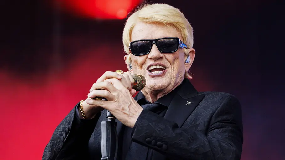 Heino hat sein Ziel erreicht. Heino singt mit Sonnenbrille und schwarzem Anzug in ein Mikrofon vor rot beleuchtetem Hintergrund.