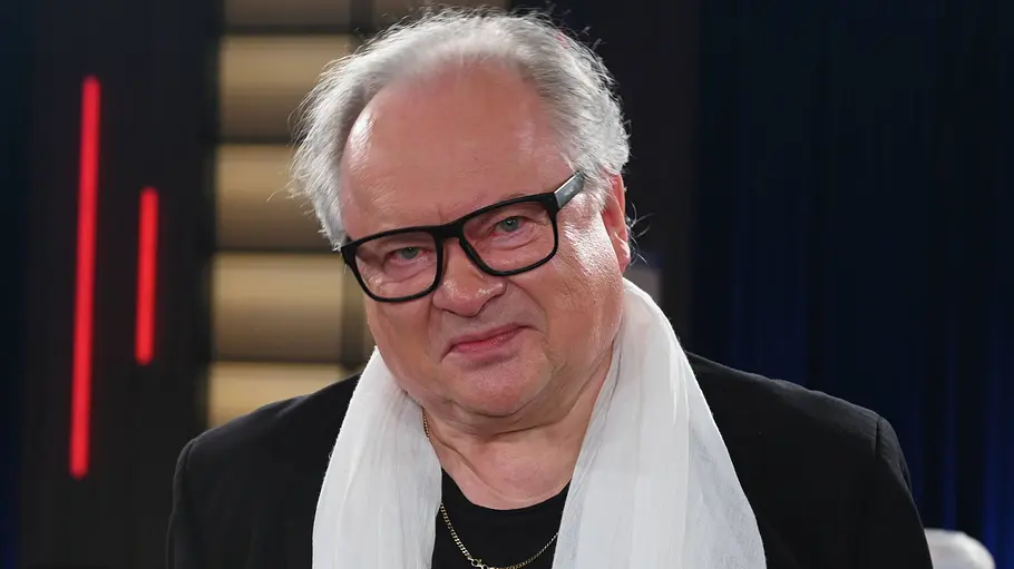 Heinz Rudolf Kunze Heinz Rudolf Kunze blickt ernst in die Kamera. Er trägt ein schwarzes Sakko, ein schwarzes T‑Shirt mit gelbem Aufdruck und einen weißen Schal. Im Hintergrund ist ein TV‑Studio mit dunklen Vorhängen, beleuchteten Wandelementen und roten Lichtstreifen zu sehen.