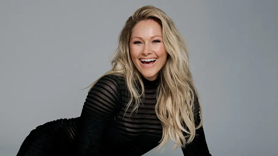 Helene Fischer, die in diesem Jahr mit ihrer „Helene Fischer Show“ zurückkehrt, liegt lächelnd in einem langen schwarzen Kleid auf grauem Boden und stützt sich mit den Händen ab.