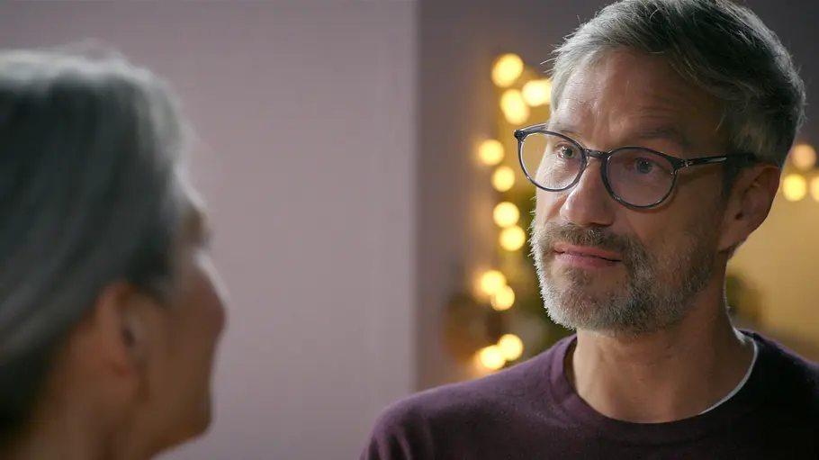Henning Daniela AWZ Henning trägt eine Brille und schaut Daniela an, die unscharf im Vordergrund steht. Im Hintergrund leuchten warme Lichter.