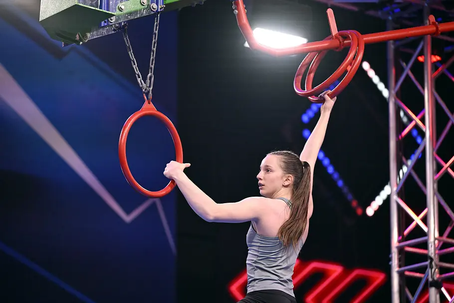 Henriette Becker ist Teilnehmer bei Ninja Warrior Germany 2026 Henriette Becker hält sich mit ausgestreckten Armen an roten Ringen eines Hindernisparcours fest. Sie trägt ein graues Top.