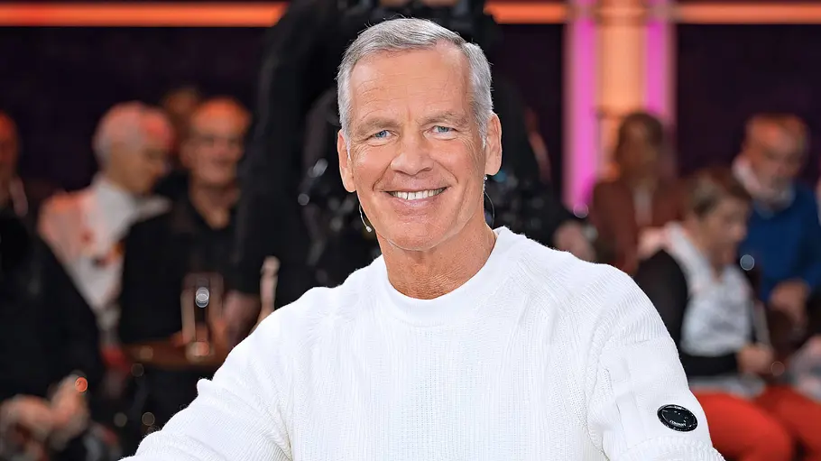 Henry Maske DAZN-Experte Henry Maske sitzt lächelnd in weißem Pullover und Jeans in einem braunen Sessel. Im Hintergrund sitzt Publikum.