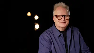 Herbert Grönemeyer mit Brille im dunklen Studio vor warmen Lichtpunkten im Hintergrund. - Foto: WDR/Daniel Waldhecker