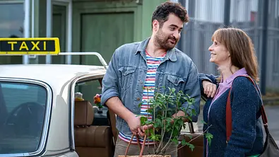 Baris und Amelie neben einem Taxi, während der Mann eine Pflanze in den Händen hält. - Foto: ARD Degeto Film/Hager Moss Film GmbH/Oliver Vaccaro Baris und Amelie neben einem Taxi, während der Mann eine Pflanze in den Händen hält. - Foto: ARD Degeto Film/Hager Moss Film GmbH/Oliver Vaccaro