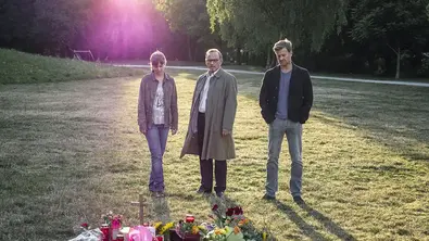 Die drei Hauptdarsteller von „München Mord“ stehen in einem Park und schauen mehrere Gedenksträuße an. - Foto: ZDF (DE) / ZDF/Hendrik Heiden