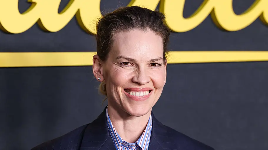 Hilary Swank lächelt in einem dunkelblauen Blazer und gestreiften Hemd vor einer dunklen Wand mit gelber Schrift.