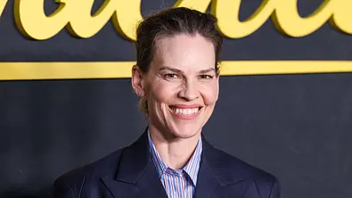 Hilary Swank lächelt in einem dunkelblauen Blazer und gestreiften Hemd vor einer dunklen Wand mit gelber Schrift. - Foto: IMAGO / Image Press Agency Hilary Swank lächelt in einem dunkelblauen Blazer und gestreiften Hemd vor einer dunklen Wand mit gelber Schrift. - Foto: IMAGO / Image Press Agency
