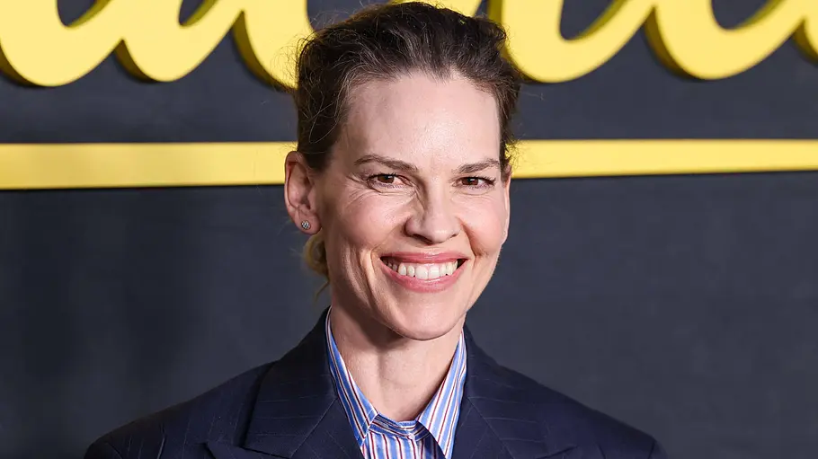 Hilary Swank drittes Kind Hilary Swank lächelt in einem dunkelblauen Blazer und gestreiften Hemd vor einer dunklen Wand mit gelber Schrift.