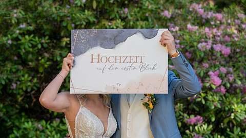 Eine Braut und ein Bräutigam halten ein Schild mit der Aufschrift Hochzeit auf den ersten Blick vor ihre Köpfe - Foto: Joyn
