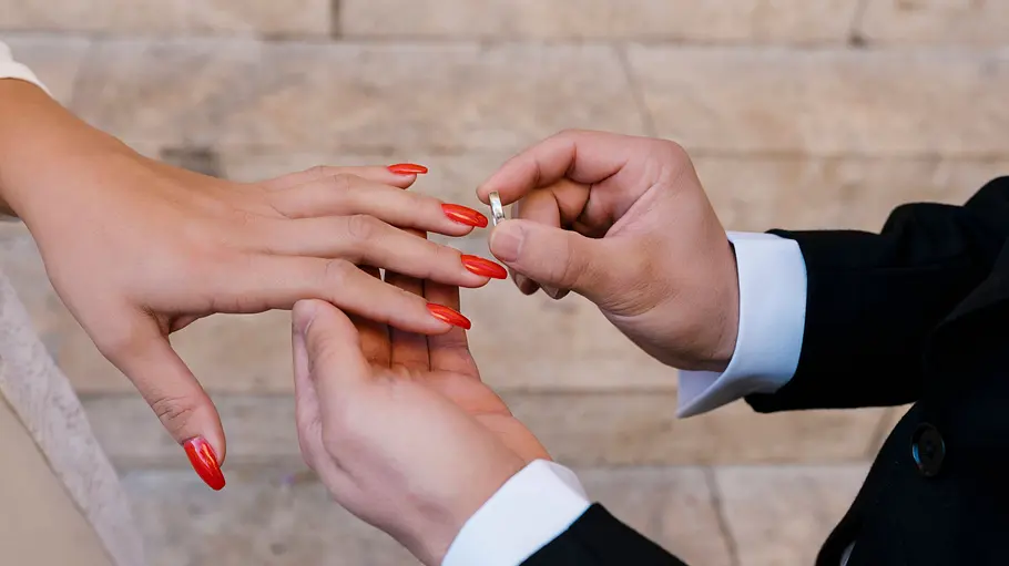 Hochzeit auf den ersten Blick geht weiter Eine Person im dunklen Anzug hält einen silberfarbenen Ring vor den Finger einer Hand mit langen, roten Fingernägeln.