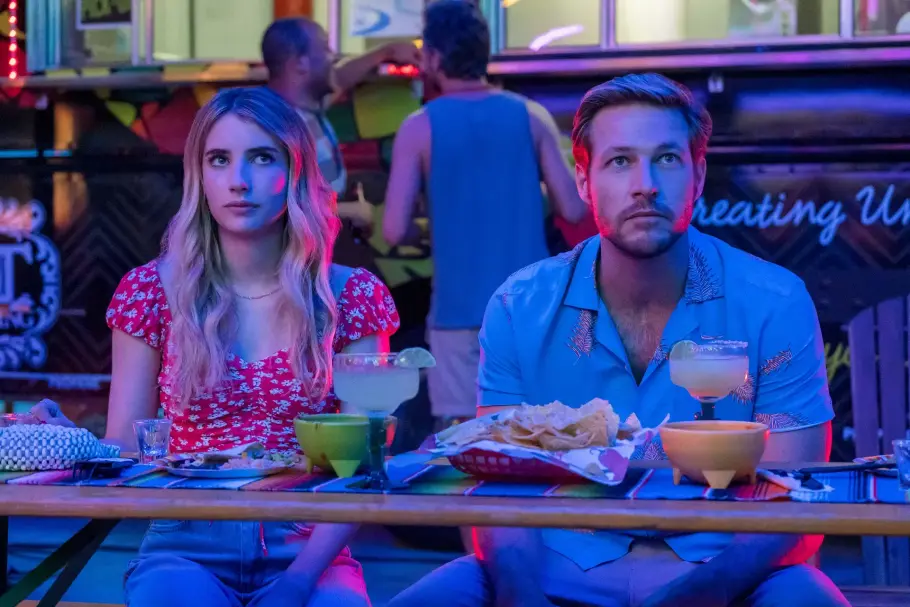Holidate Ausschnitt aus dem Film Holidate mit Emma Roberts als Sloane Reed und Luke Bracey als Jackson. Sie sitzen nebeneinander. Vor ihnen stehen Cocktails und Nachos auf dem Tisch.