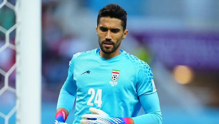 Torhüter der iranischen Nationalmannschaft im hellblauen Trikot und mit Torwarthandschuhen vor dem eigenen Tor. Er blickt konzentriert nach unten, während der Hintergrund des Stadions unscharf ist.