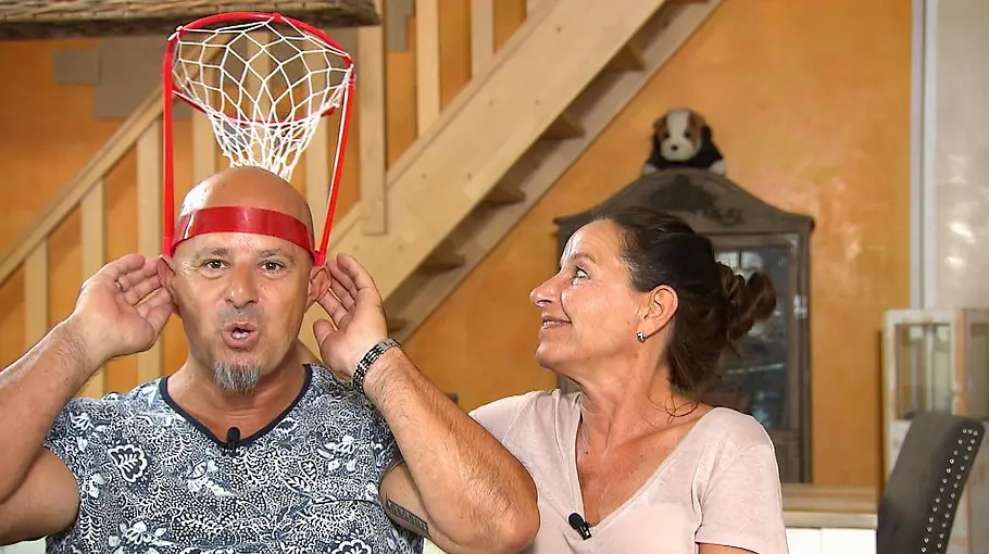 Detlef und Nicole Steves Ein Mann trägt ein kleines Basketballkorb-Spiel auf dem Kopf und hält es mit beiden Händen fest, während eine Frau neben ihm sitzt und ihn lächelnd anschaut; im Hintergrund ist ein Wohnraum mit Treppe und Möbeln zu sehen.