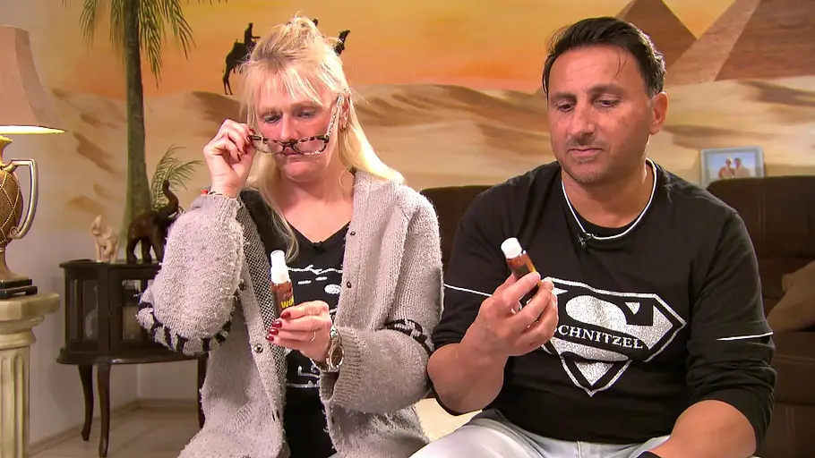 Ehepaar Bartsch Ein Mann und seine Frau sitzen auf einem Sofa und begutachten kleine Fläschchen; die Frau hebt ihre Brille an, während im Hintergrund eine Wüstenlandschaft mit Pyramiden als Wandmotiv zu sehen ist.