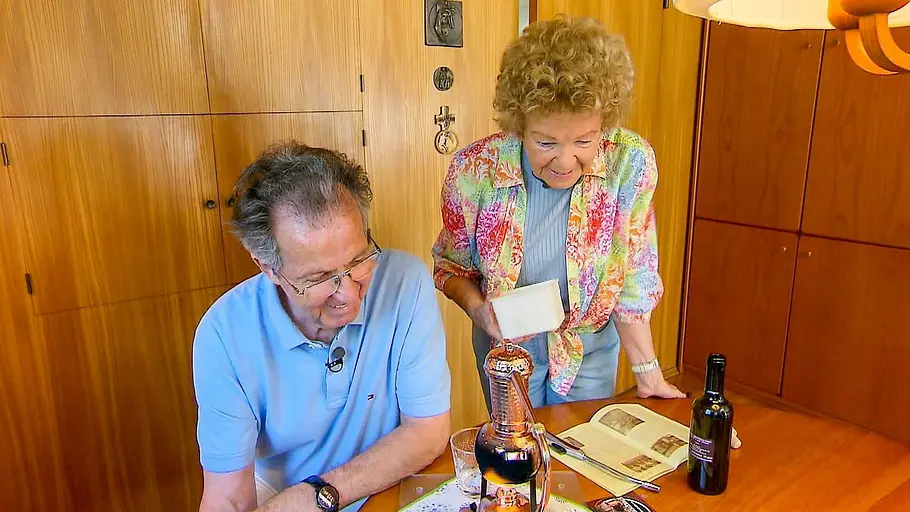 Ingrid und Otto Kneidinger Ein älteres Paar steht an einem Holztisch in einem wohnlichen Raum; der Mann sitzt und schaut auf einen Gegenstand auf dem Tisch, während die Frau neben ihm eine kleine Schachtel in der Hand hält.