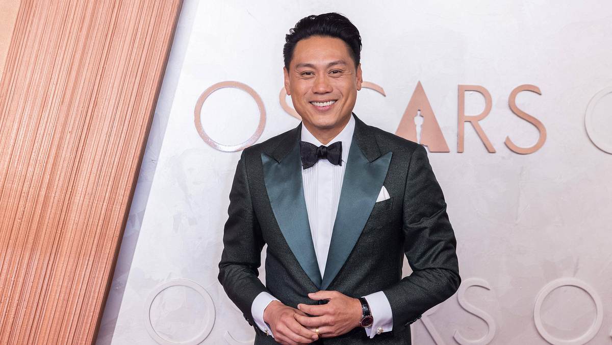 Hot Wheels Jon M. Chu ist vor einer Fotowand der Oscars. Er trägt einen schwarzen Anzug mit einer schwarzen Fliege.