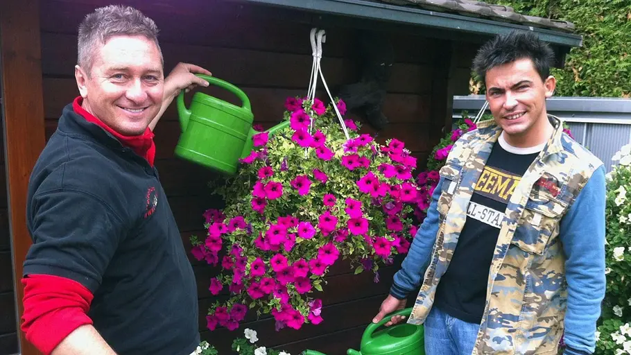 Hubert und Matthias Zwei Männer stehen vor einem Gartenhaus und gießen mit grünen Gießkannen üppig blühende Hängepetunien in Pink und Weiß.