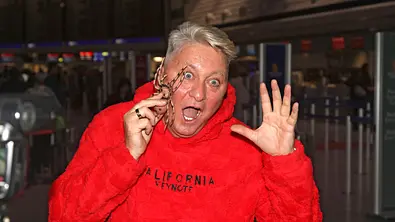 Hubert Fella ist am Flughafen, er hält eine Plastikspinne in der Hand und tut erschrocken. - Foto: IMAGO / Future Image