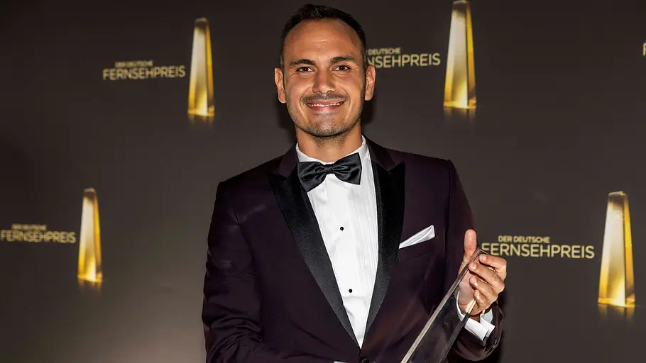 Ibrahim Naber Ein lächelnder Mann im dunklen Smoking mit Fliege hält eine transparente Trophäe vor einer Wand mit Logos des Deutschen Fernsehpreises.