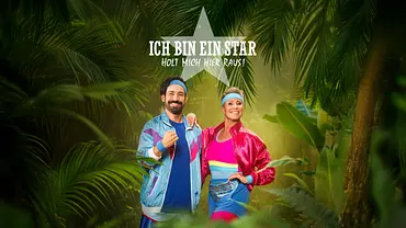 Zwei gut gelaunte Moderatoren in bunten Retro-Trainingsanzügen und Stirnbändern stehen lächelnd inmitten eines dschungelartigen Hintergrunds. Über ihnen prangt ein großer Stern mit dem Schriftzug „Ich bin ein Star – Holt mich hier raus!“. - Foto: RTL / Pascal Bünning